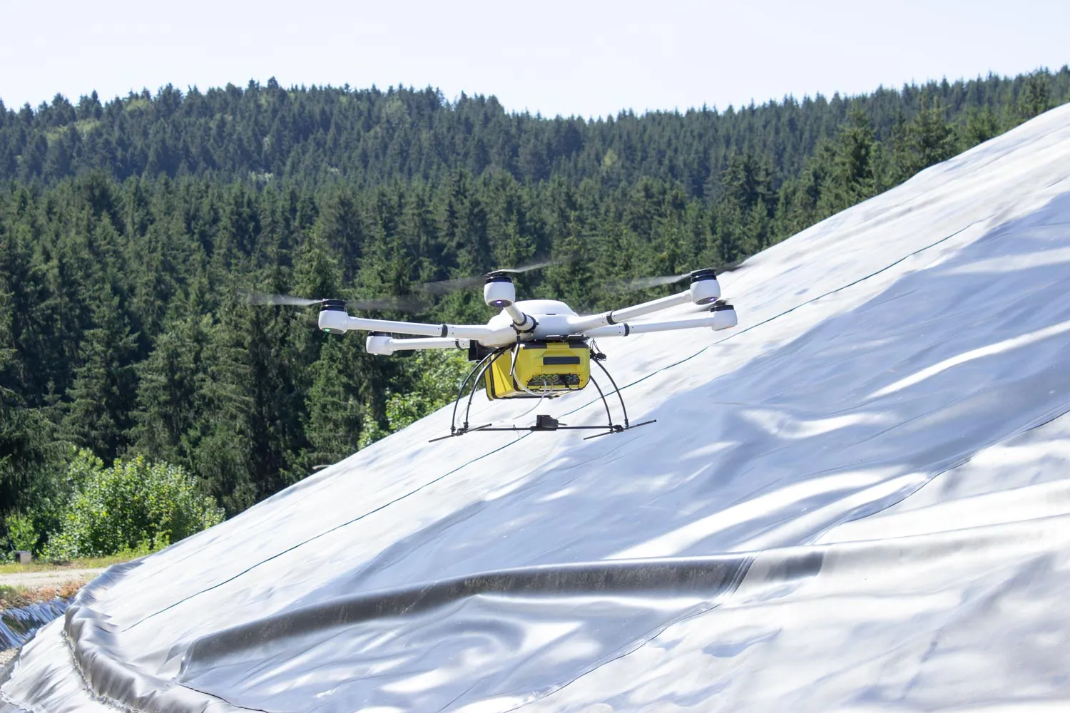 Exabotix Atlas Drohne mit schwerem Gas-Sensor während eines Inspektionsfluges über eine mit einer Plane abgedeckten Fläche in einem Waldgebiet.